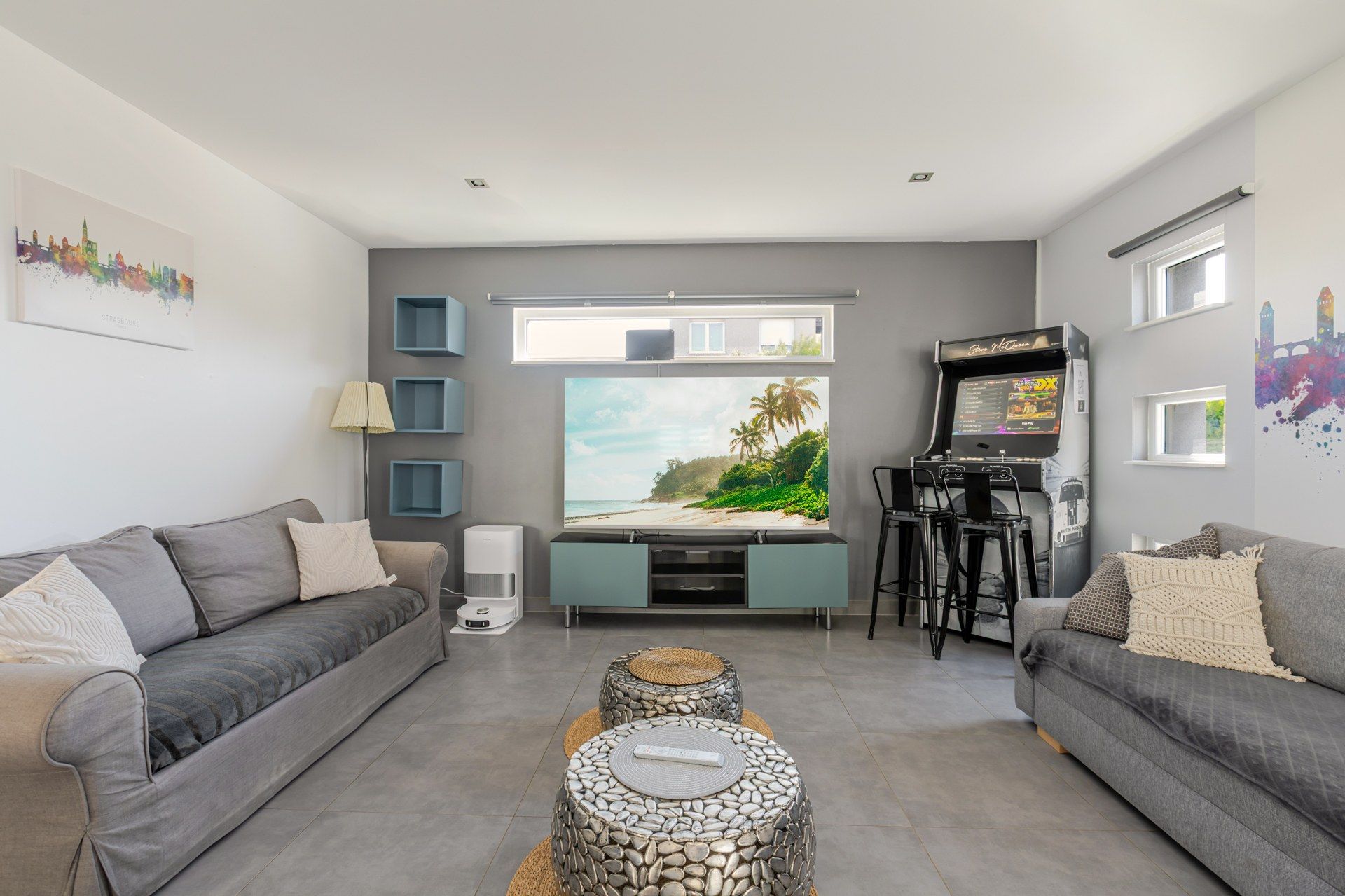 A living room with grey sofas, a flat-screen TV, a gaming console, and a large window with a beach view.