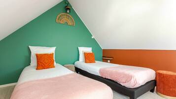 A room with two beds, one with a black frame and the other with a white frame, both with pink bedding. There is a small round orange ottoman and a wall-mounted light fixture.