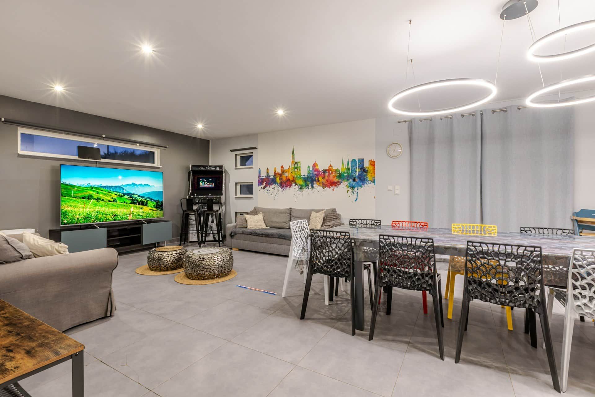 A modern living room with a flat-screen TV, a gaming console, a sofa, a dining table with chairs, and a colorful cityscape mural.