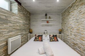 A bedroom with a stone wall, a bed with floral pillows, a bedside table with a lamp, and a shelf with decorative items. - Villa 250m² with private spa, barbecue, table football (Hurtigheim)