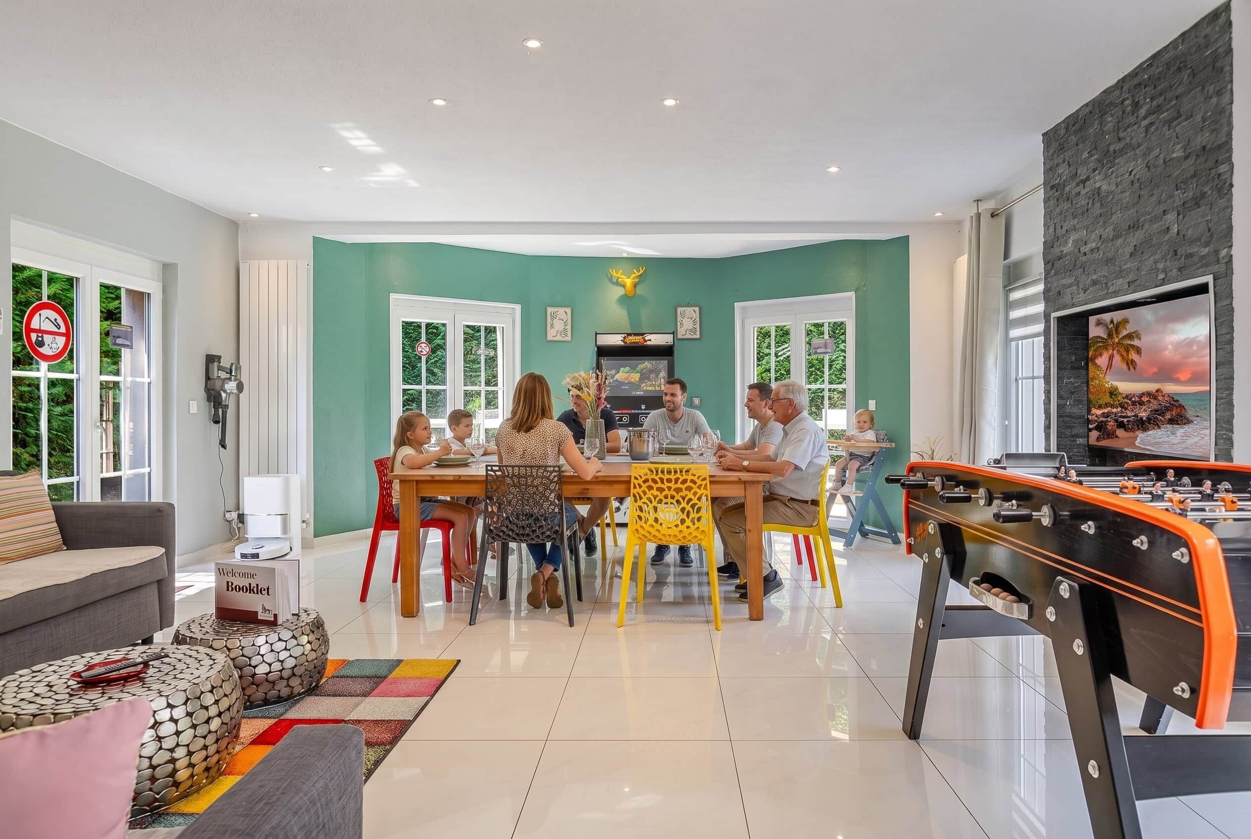 Un área de comedor con un grupo de personas sentadas alrededor de una mesa, una mesa de futbolín y un televisor mostrando una vista panorámica.
