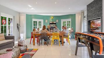 Un área de comedor con un grupo de personas sentadas alrededor de una mesa, una mesa de futbolín y un televisor mostrando una vista panorámica.