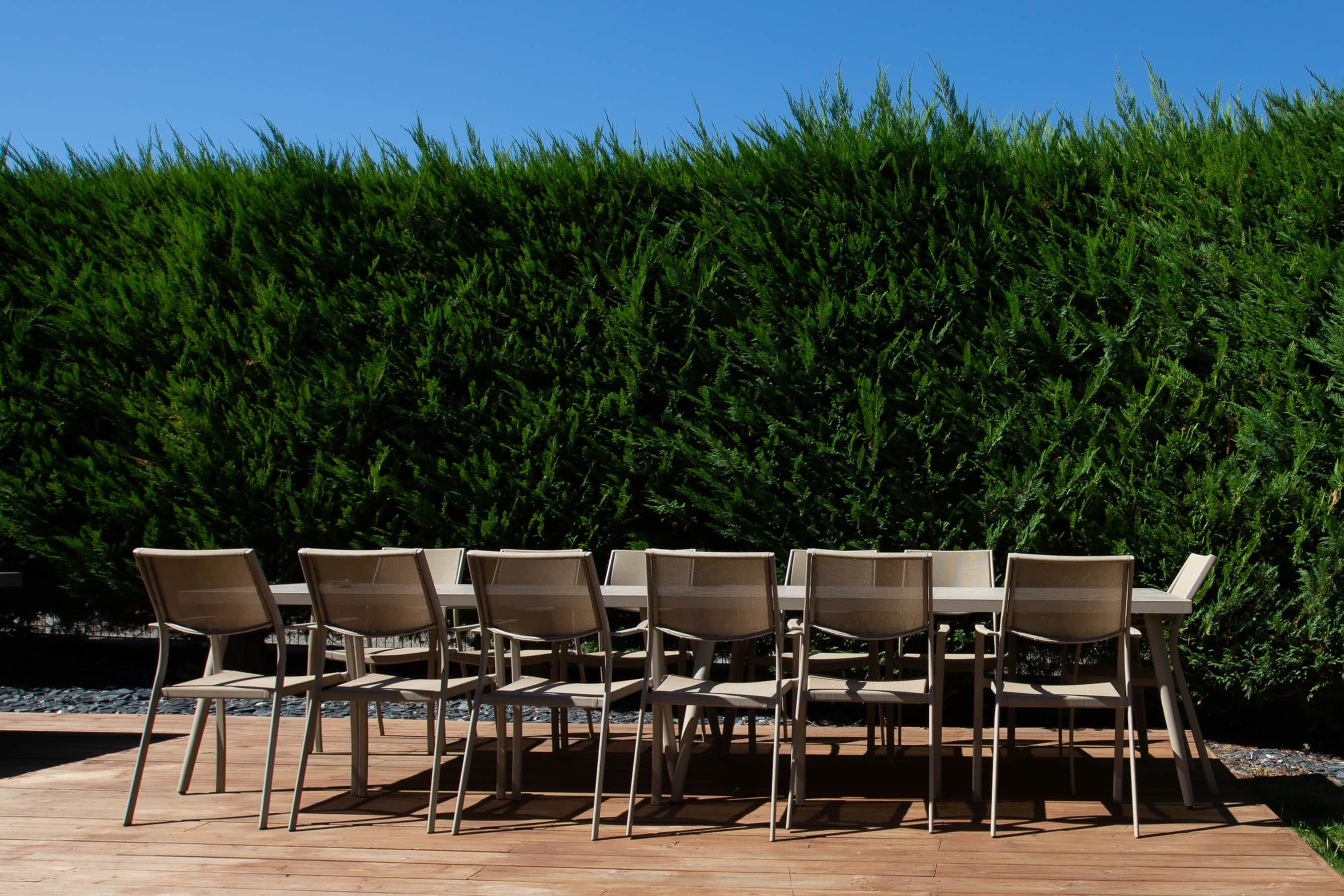 An outdoor dining area with six beige chairs and tables, set against a backdrop of dense green foliage.