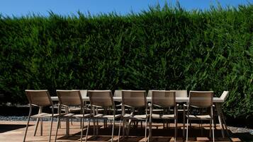 An outdoor dining area with six beige chairs and tables, set against a backdrop of dense green foliage.