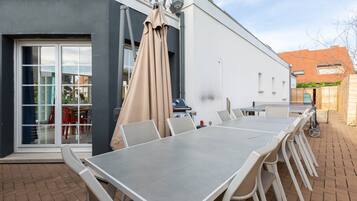 An outdoor dining area with a long table and chairs, a covered seating area, and a building entrance with glass doors.