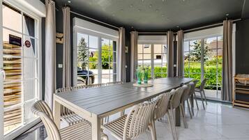 Un espace repas avec une longue table et des chaises, de grandes fenêtres et une vue sur la verdure environnante.