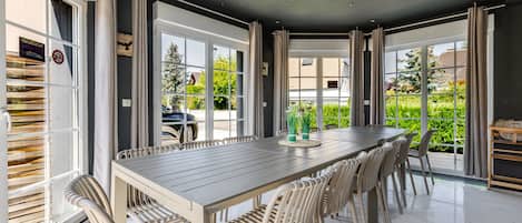 A dining area with a long table and chairs, large windows, and a view of greenery outside.
