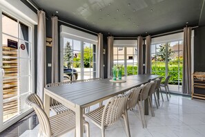 A dining area with a long table and chairs, large windows, and a view of greenery outside.