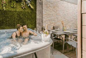 A hot tub with a couple, a table with chairs, and a brick wall with greenery. - Gites Spa Strasbourg Appartement le 14 jacuzzi sauna privé, Gite le 14 (Furdenheim)