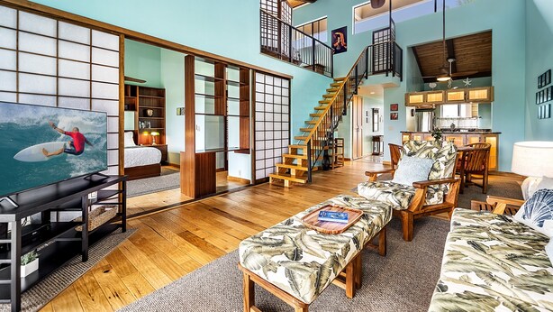 A spacious living area with a wooden floor, a large flat-screen TV, a sofa, and a dining area with wooden furniture. - KKSR#7303 Remodeled Top Floor Unit With Loft, Golf Course Views (Kailua Kona)