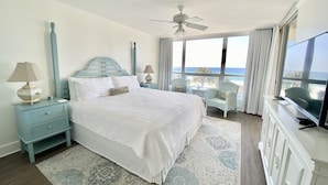A bedroom with a bed, nightstands, a ceiling fan, a TV, a view of the ocean, and a balcony with chairs.