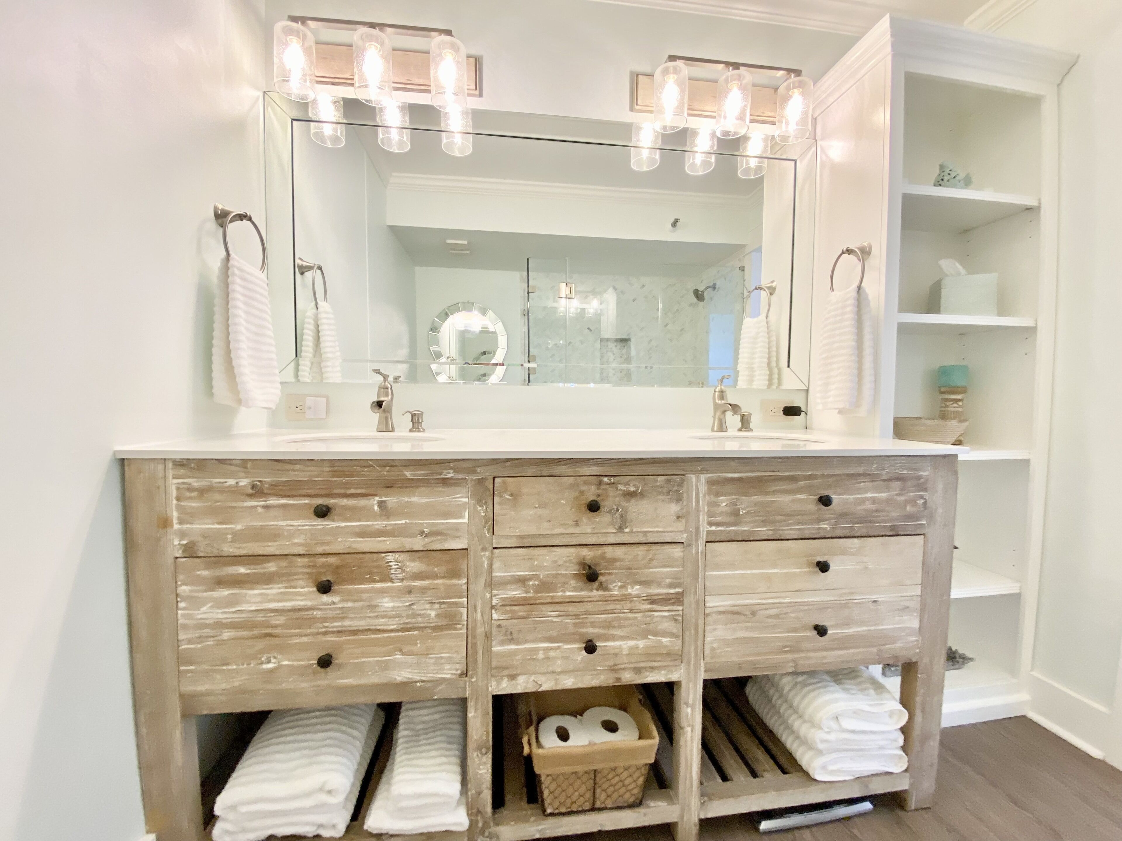 Une salle de bain avec un meuble-lavabo blanc, deux vasques et un grand miroir avec éclairage. On y trouve des serviettes et un panier placé sous le meuble-lavabo.