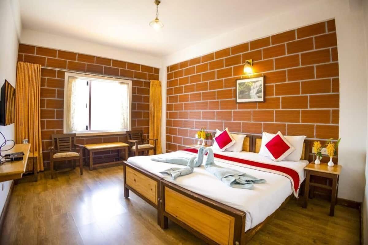 A bedroom with a brick wall, a bed with pillows, a bedside table with flowers, a window with curtains, and a desk with a chair.