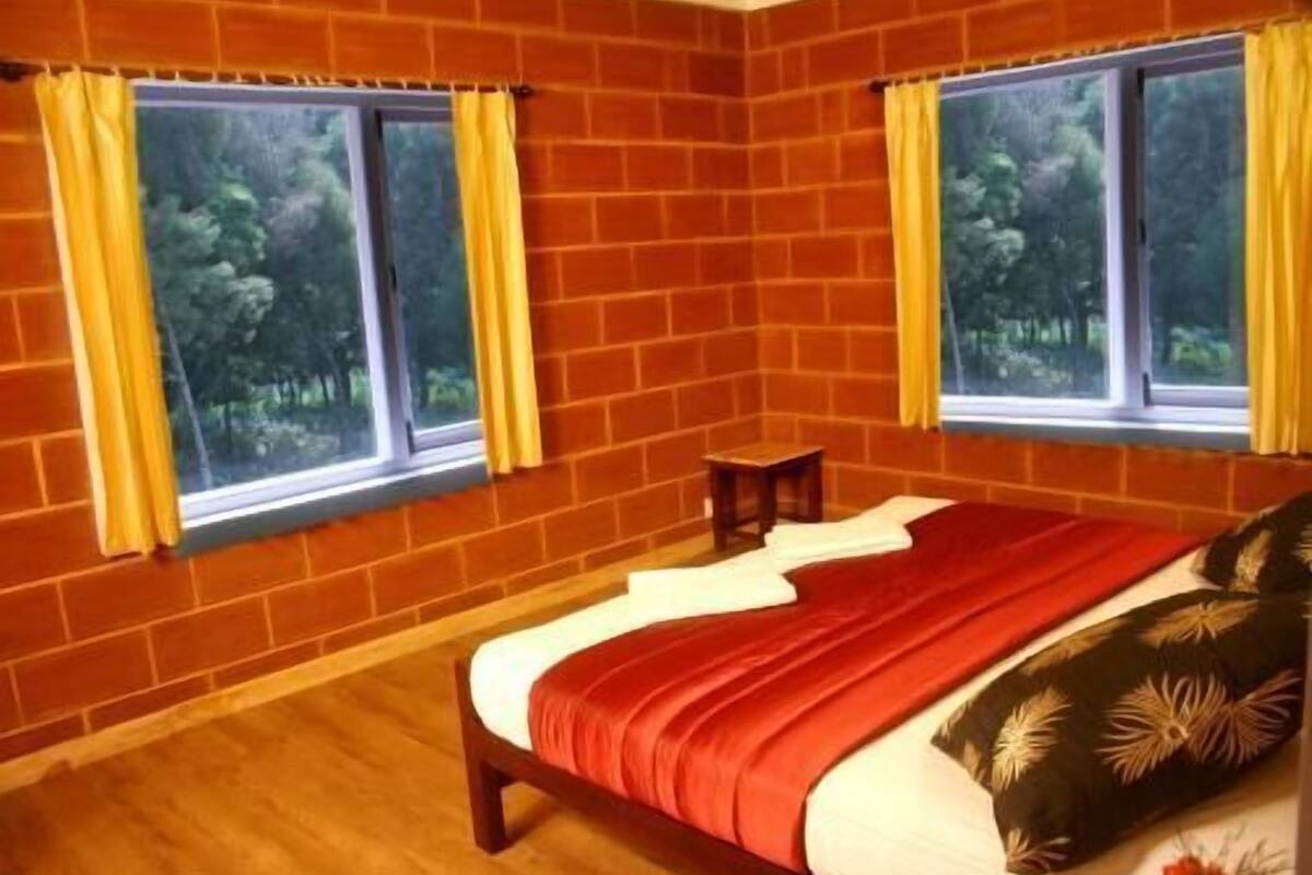 A bedroom with a brick wall, two windows with curtains, a bed with a red blanket, and a small wooden table.