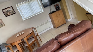 Una sala de estar con un sofá de cuero marrón, una mesa de comedor de madera con sillas, un televisor montado en la pared y una ventana con persianas.