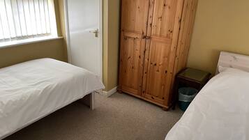A bedroom with two single beds, a wooden wardrobe, a small bedside table, and a window with blinds.