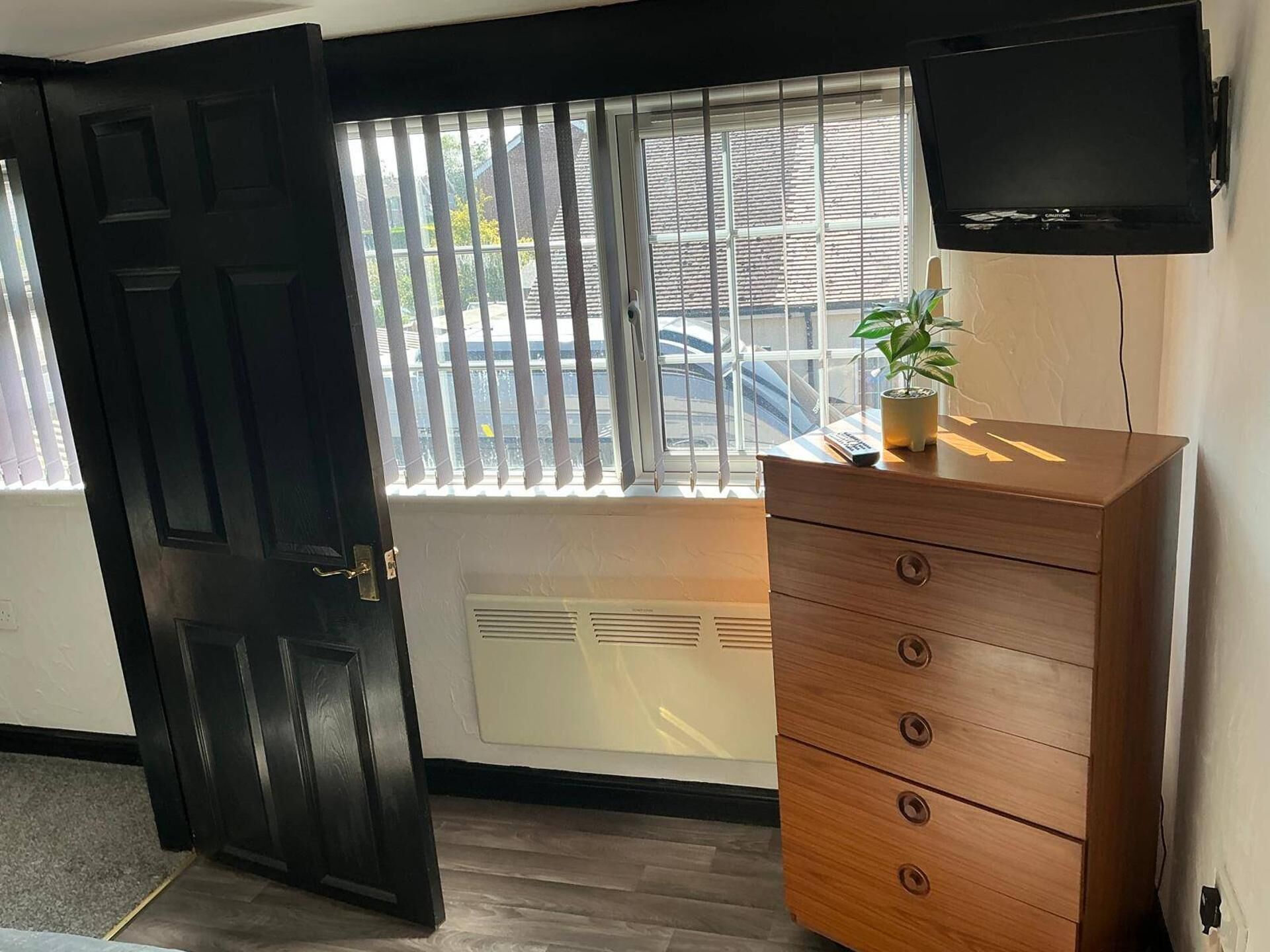 A room with a wooden dresser, a television mounted on the wall, a window with blinds, and a door.