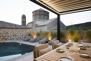 Un espace de restauration aménagé au bord de la piscine, avec une table dressée pour deux, donnant sur un bâtiment en pierre doté d’un clocher.