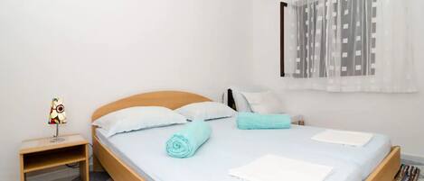 A neatly made bed with white linens and a blue towel, a bedside table with a lamp, and a window with sheer curtains.