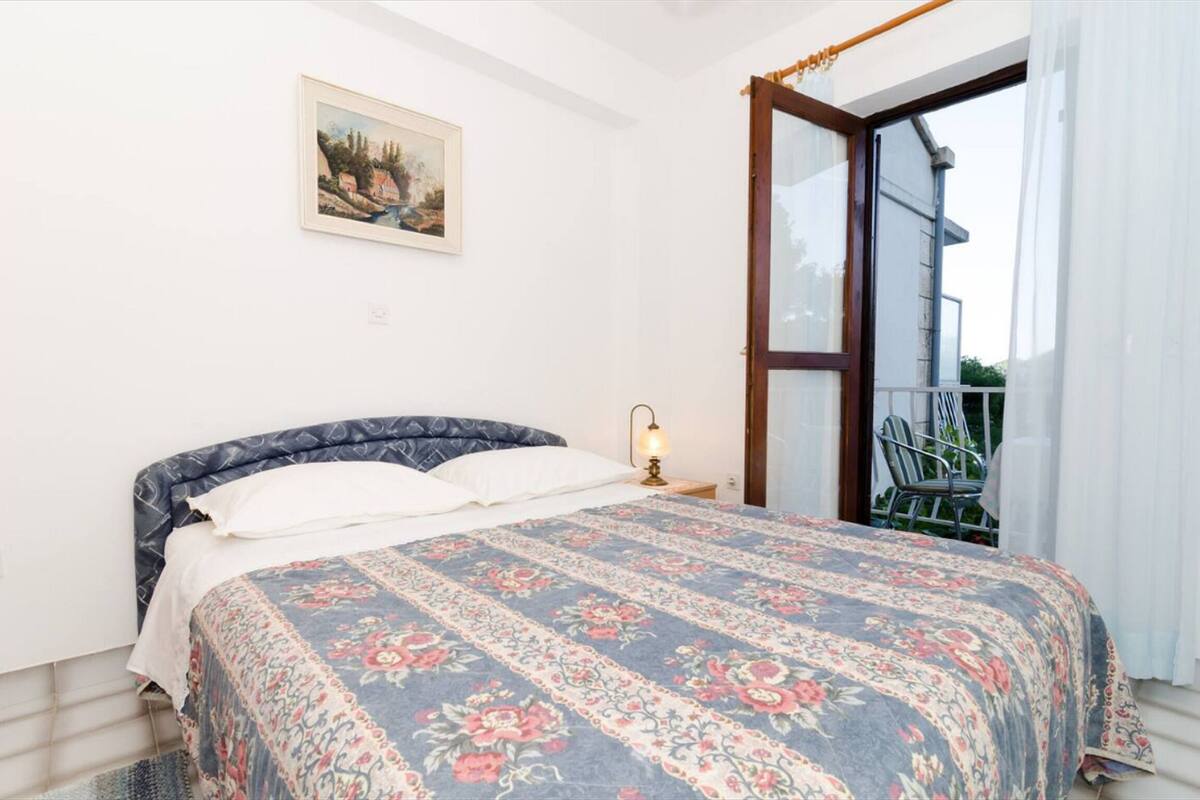 Ein Schlafzimmer mit einem Bett, einem Nachttisch, einer Lampe, einem gerahmten Bild an der Wand und einem Balkon mit einem Stuhl.