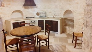 A rustic stone kitchen with a round wooden table and chairs, a built-in oven, and a hanging light fixture. - Trulli in Monopolitan countryside  (Puglia)