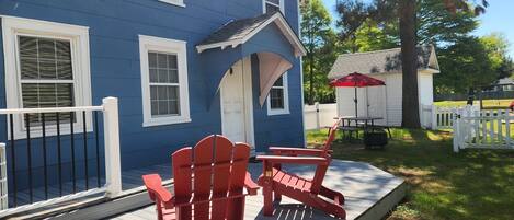 Une maison bleue avec un porche, deux chaises Adirondack rouges, un petit abri de jardin et une clôture blanche en piquets.