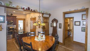 A dining area with a wooden table and chairs, a chandelier, and a kitchen visible through an open door.