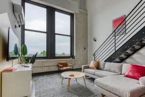 A modern living room with a large window, a sofa, a coffee table, and a staircase. - Sosuite | 2BR Loft w W/D, Gym, Lounge (Philadelphia)