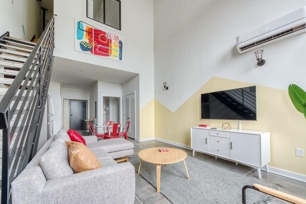 A spacious living area with a grey sofa, a round wooden coffee table, and a white cabinet with a flat-screen TV. A staircase is visible in the background, and an air conditioning unit is mounted on the wall. - Sosuite | 2BR Loft w W/D, Gym, Lounge (Philadelphia)