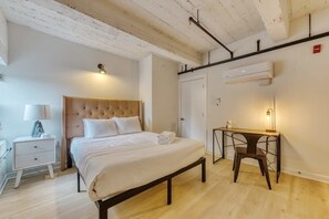 A bedroom with a bed, bedside table, lamp, desk, chair, and an air conditioning unit. - Sosuite | 2BR Loft w W/D, Gym, Lounge (Philadelphia)