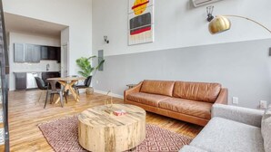 A modern living room with a brown leather sofa, a round coffee table, and a grey armchair. There is a dining area with a wooden table and chairs, and a kitchen in the background. - Sosuite | 2BR Loft w W/D, Gym, Lounge (Philadelphia)