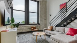 A modern living room with a large window, a sofa, a coffee table, and a staircase. - Sosuite | 2BR Loft w W/D, Gym, Lounge (Philadelphia)