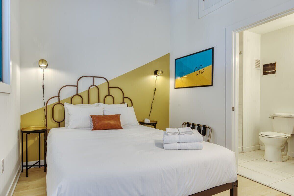 A neatly made bed with white linens and a brown pillow, a bedside table with a lamp, and a painting on the wall.