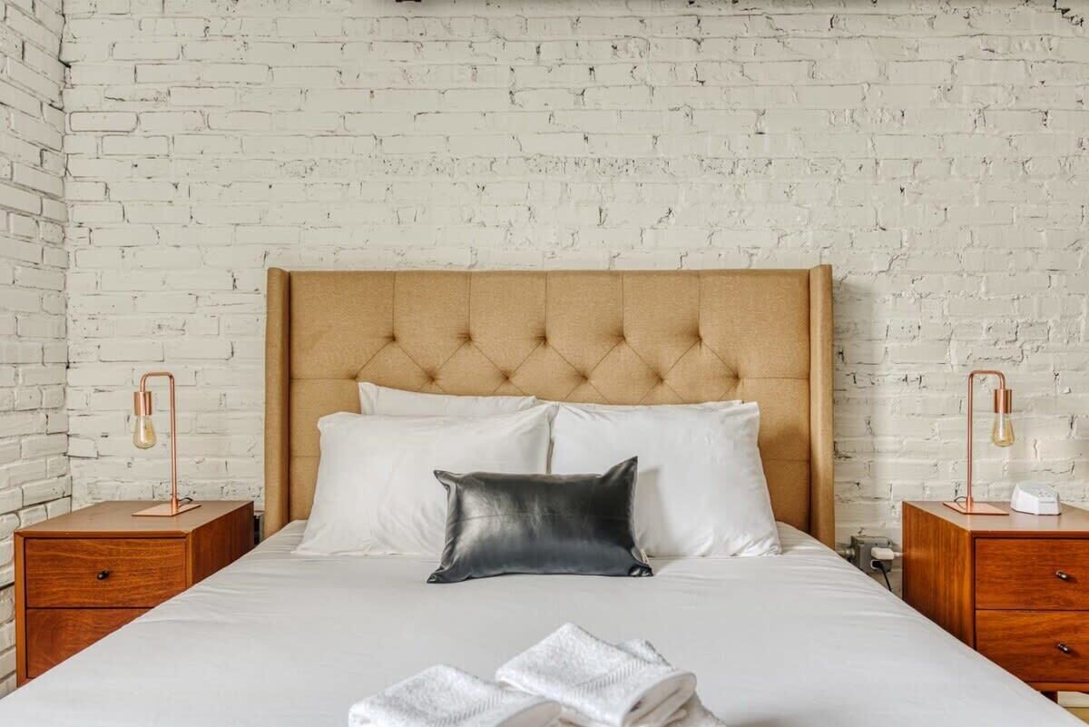A neatly made bed with a tufted headboard, two bedside tables with lamps, and a white brick wall.