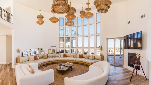 A spacious living room with a curved sofa, a coffee table, and a fireplace. There are large windows providing natural light and a view of the outdoors. - Casa Blanca by Fieldtrip - Boulder Estate with Hot Tub, Soaking Pool & Outdoor Shower (Yucca Valley)