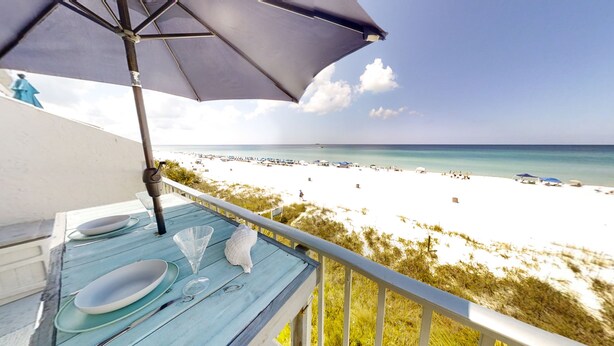 A balcony with a view of a beach, a table set with plates and a glass, and an umbrella. - Gulf front 2 bedroom with stunning views & Free round of Golf Daily! (Panama City Beach)