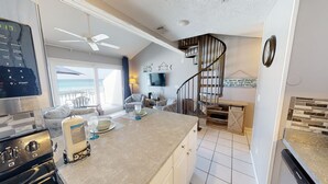 A kitchen with a modern stove, a dining area with a table and chairs, a TV mounted on the wall, and a staircase leading to a balcony with a view of the ocean. - Gulf front 2 bedroom with stunning views & Free round of Golf Daily! (Panama City Beach)