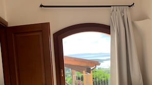 A room with a wooden door, a window with a view of a balcony and outdoor scenery, and a desk with drawers. - Villa Conca Verde (Conca Verde (SS))