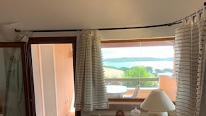 A room with a view of a body of water, a wooden cabinet, a table with a lamp, and a window with curtains. - Villa Conca Verde (Conca Verde (SS))