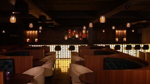A dimly lit restaurant with wooden booths and tables, a bar area with hexagonal light fixtures, and a patterned wall in the background.