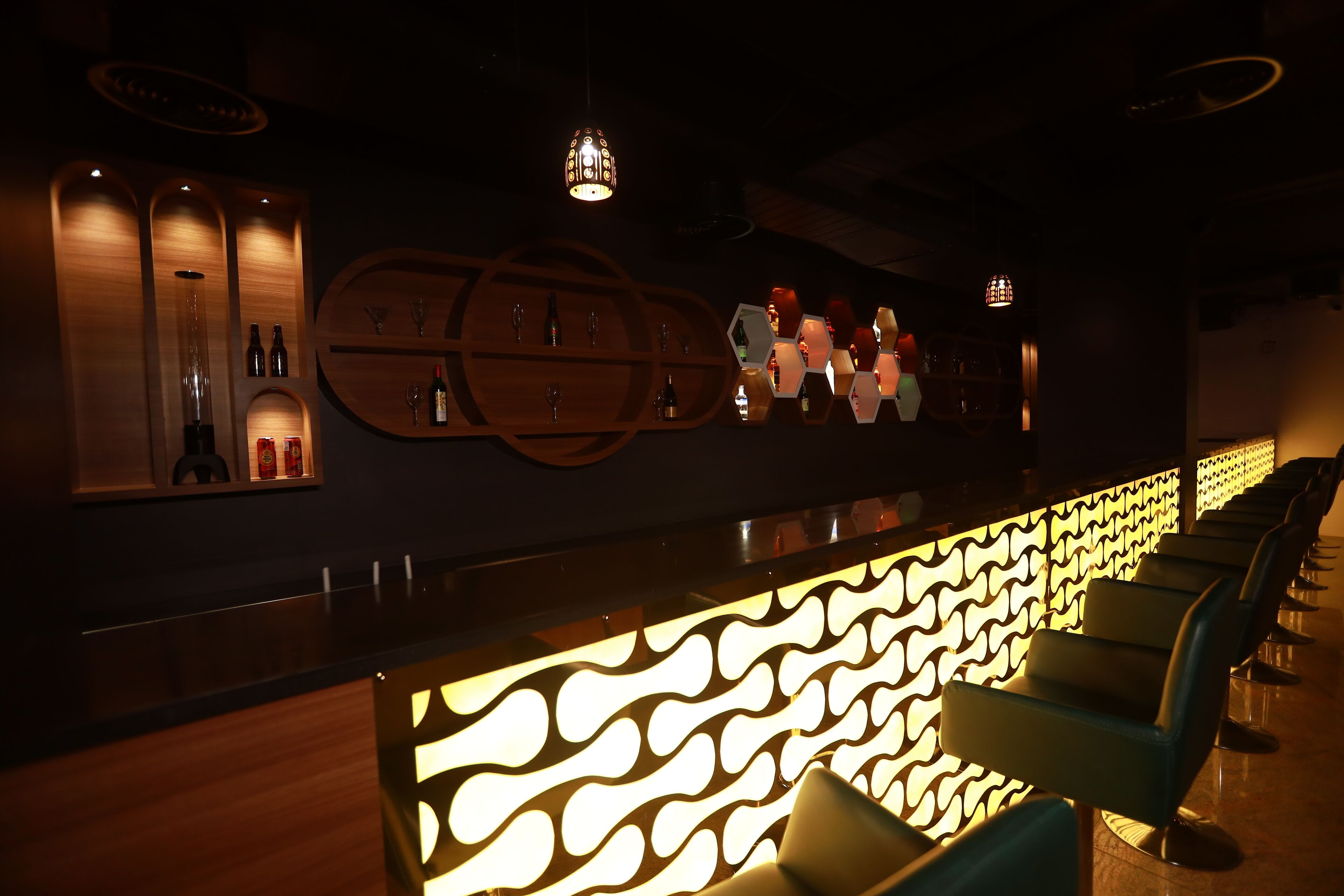 Zona de bar con mostrador de madera, estantes con botellas y copas, y pared decorada con un diseño luminoso.