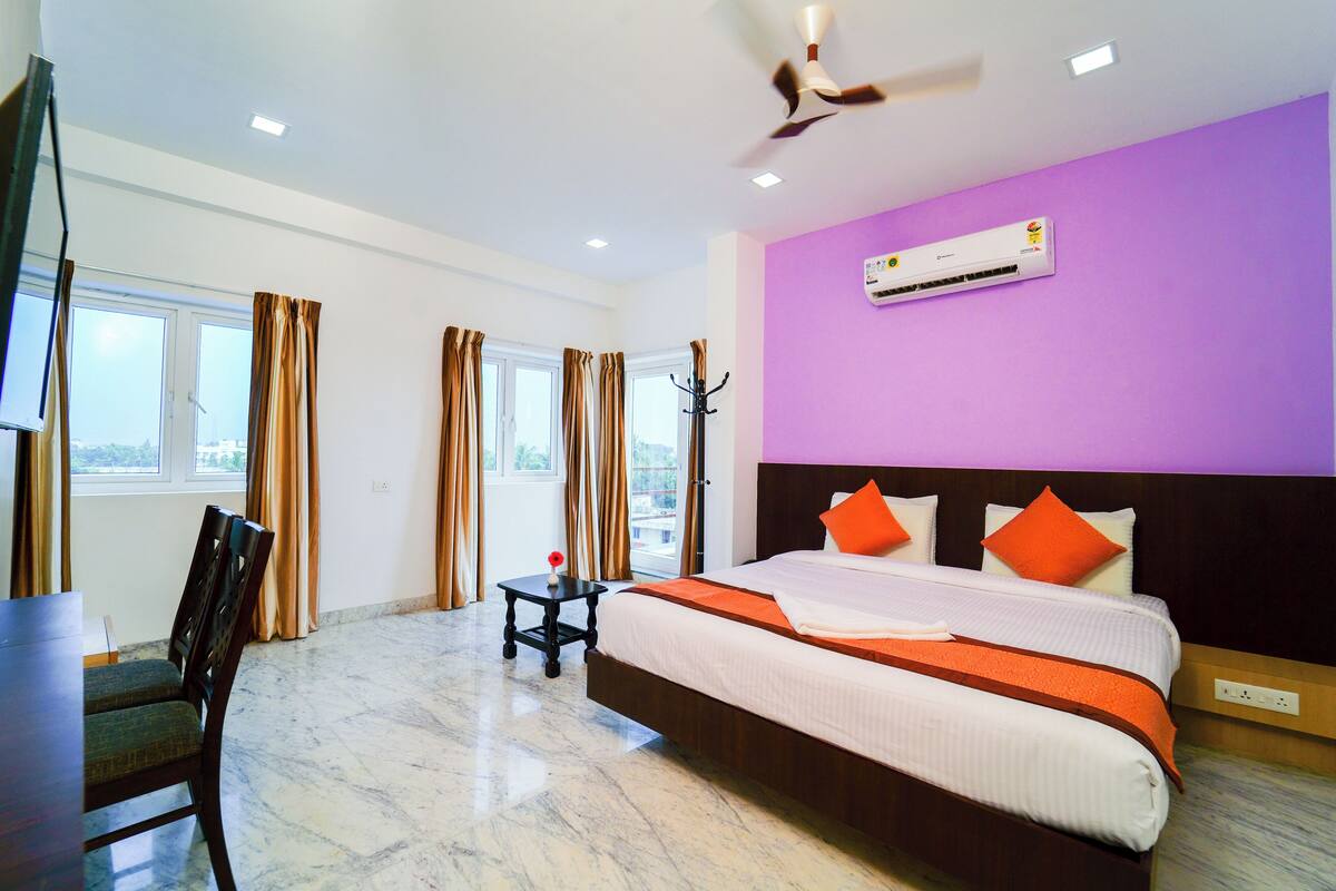 Une chambre d’hôtel avec un grand lit, un bureau et une chaise. On y trouve un ventilateur de plafond, un appareil de climatisation et une fenêtre avec des rideaux.