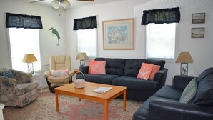 A living room with a sofa, armchair, and a coffee table. There are two lamps, a framed picture on the wall, and a window with curtains.