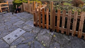 Une terrasse pavée de pierres, avec une clôture en bois, un regard d’égout et une vue sur des collines ondoyantes et un ciel nuageux.