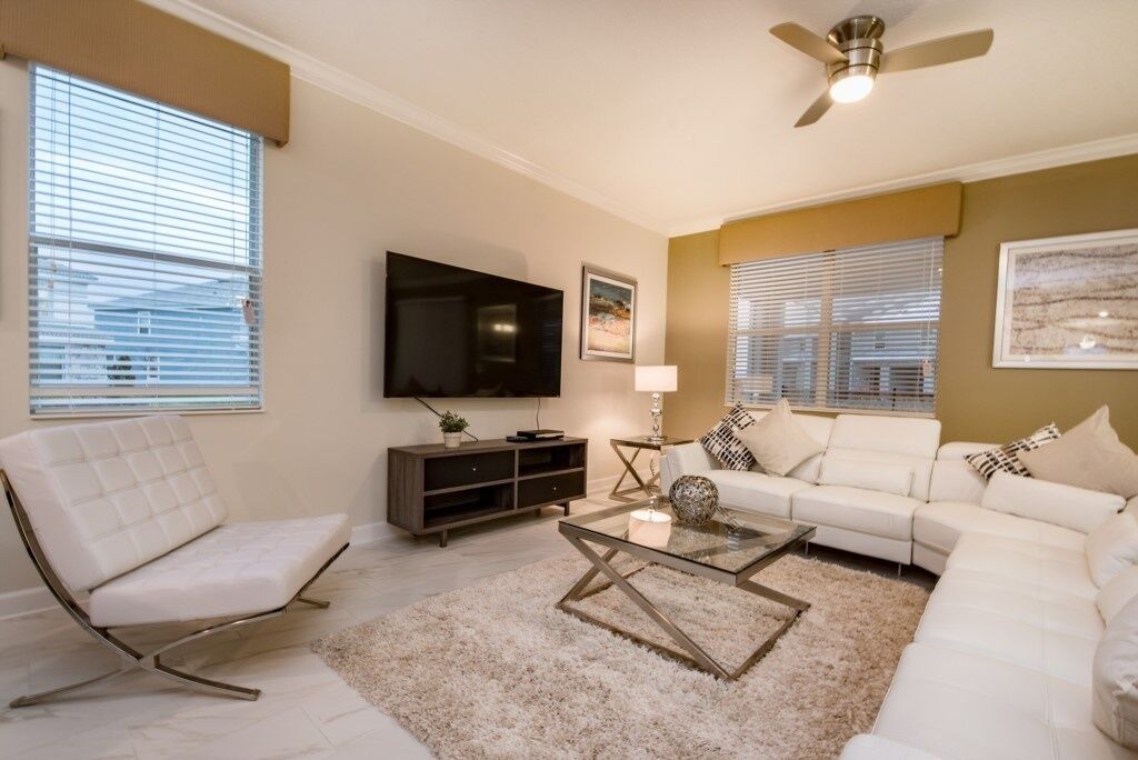 A living room with a white sofa, a white armchair, a glass coffee table, a TV on the wall, and a ceiling fan.
