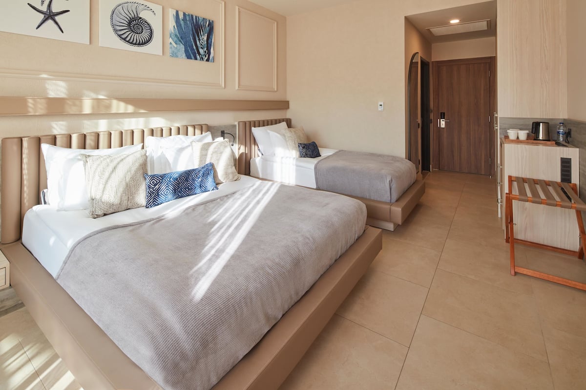 A hotel room with two beds, a wooden headboard, a small kitchenette, and a door leading to another room.