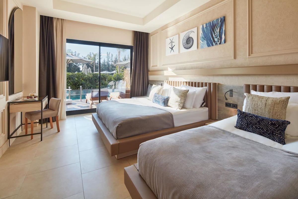 A hotel room with two beds, a desk, and a chair. There is a large window with a view of the outdoors.