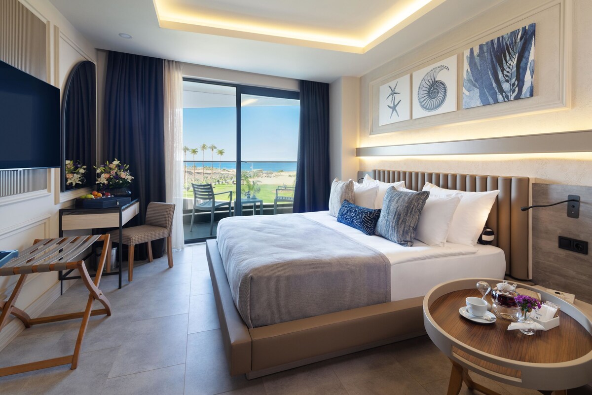 Una habitación de hotel con una cama grande, un escritorio con una silla, una mesita con una taza y flores, y un balcón con vista al océano.