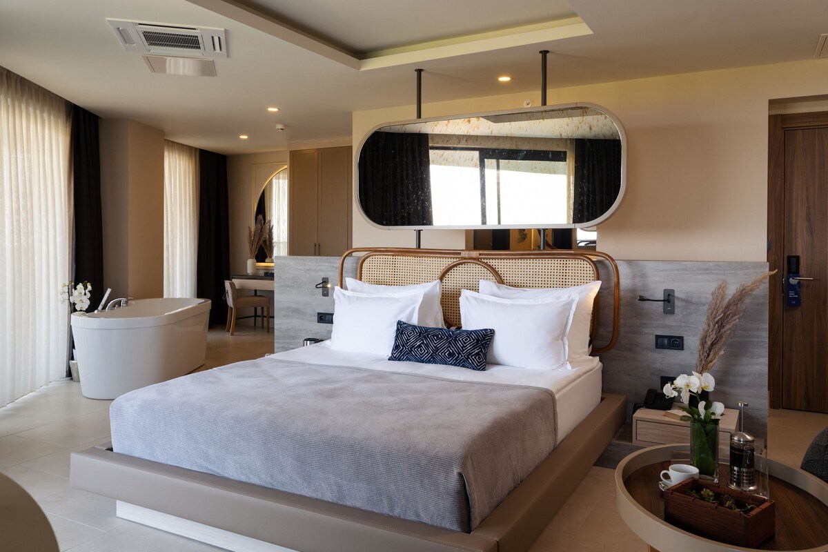 Habitación de hotel moderna con una cama grande, una zona de estar con una mesita pequeña, una bañera y un espejo grande sobre la cama.