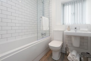 Un baño moderno con azulejos blancos, ducha con mampara de vidrio, inodoro blanco y lavabo con espejo arriba.